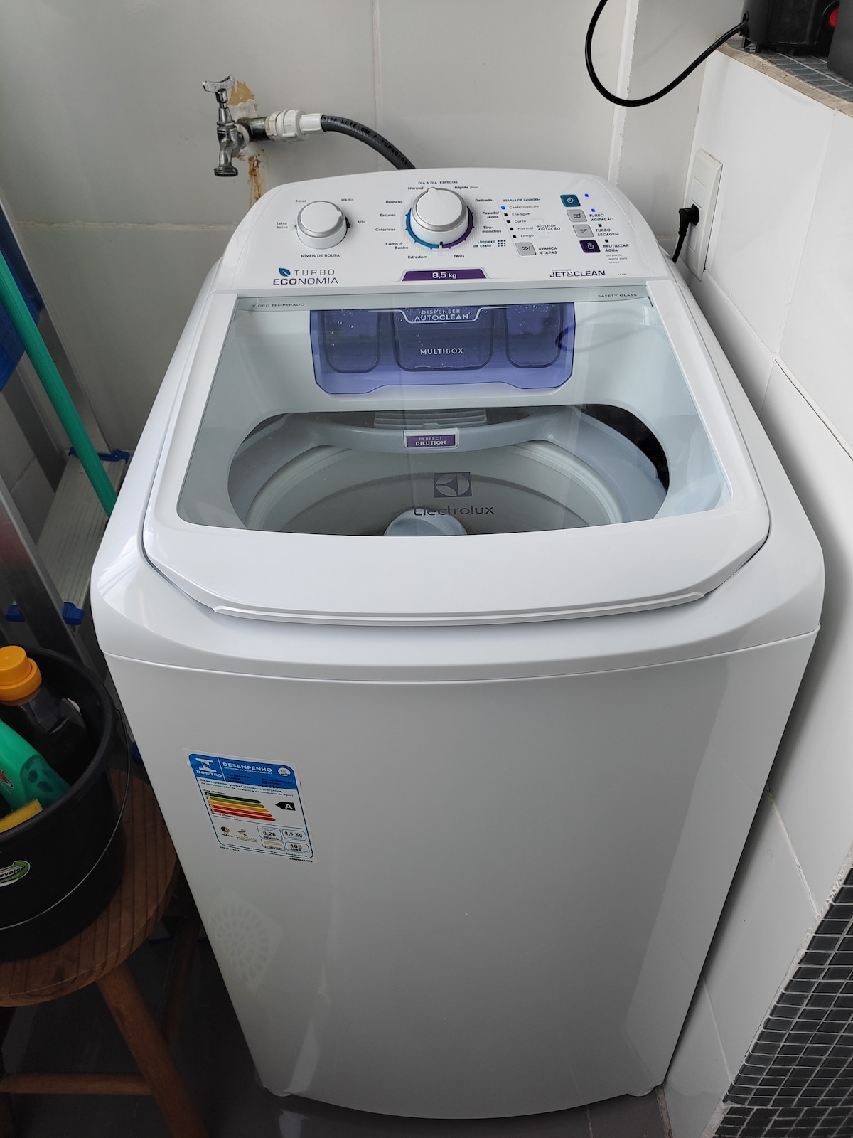 A modern washing machine is positioned in a laundry area, featuring a clear lid that provides visibility of the interior. Control dials and settings are located on the top, with a visible detergent compartment. A wooden stool and cleaning supplies are nearby.