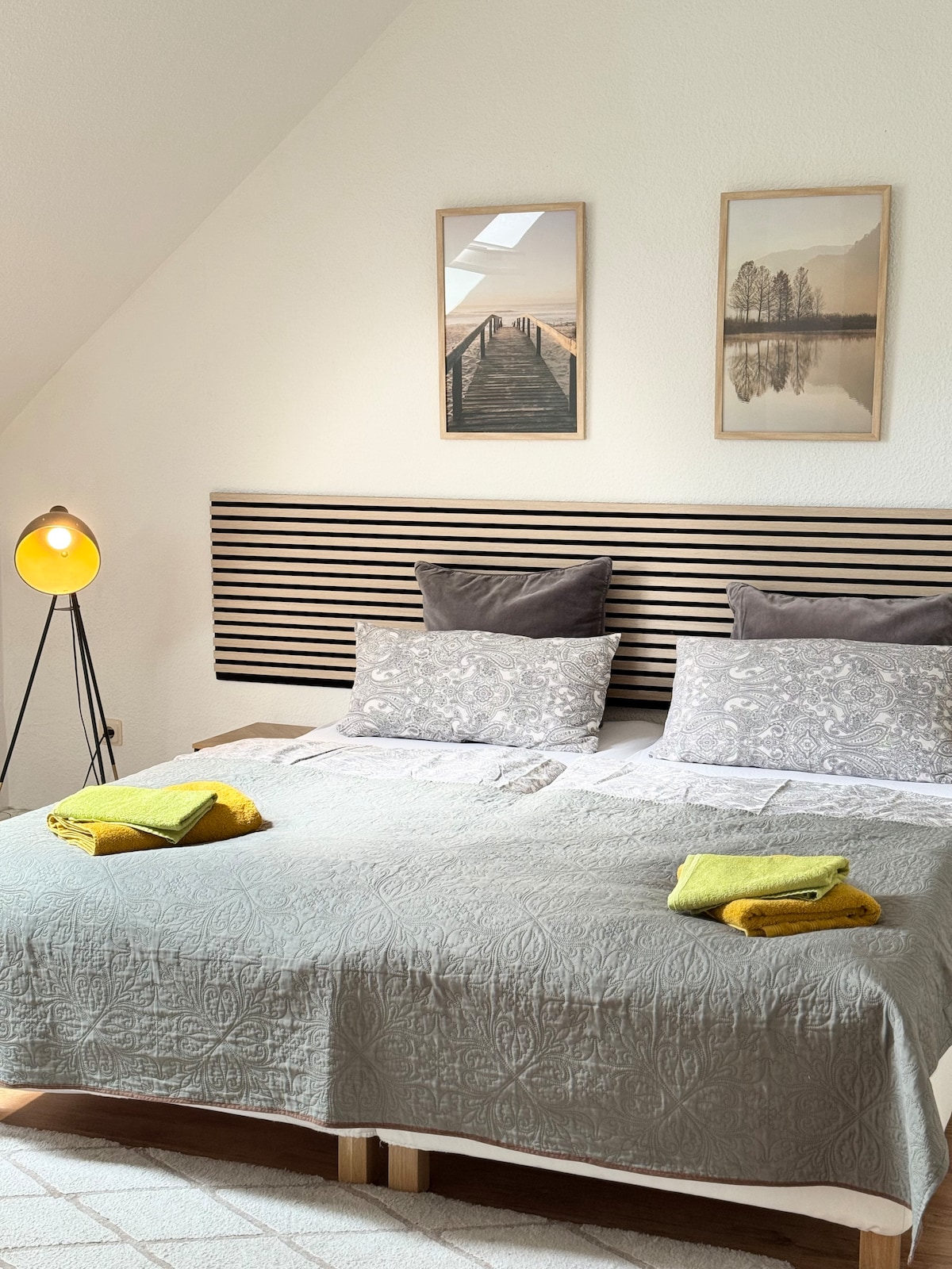 A spacious sleeping area is highlighted by a bed with a grey patterned coverlet and two decorative pillows. Warm light from a standing lamp enhances the space, while framed artwork featuring a natural landscape hangs on the wall. Fresh towels are neatly arranged on the bed.