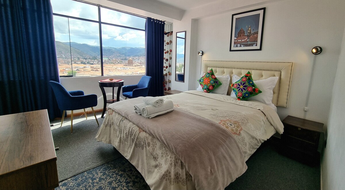A spacious bedroom is highlighted by a large window with views of the surrounding landscape. A well-made bed with decorative pillows is positioned near a cozy seating area featuring two armchairs and a small table. Soft lighting complements the tranquil environment.