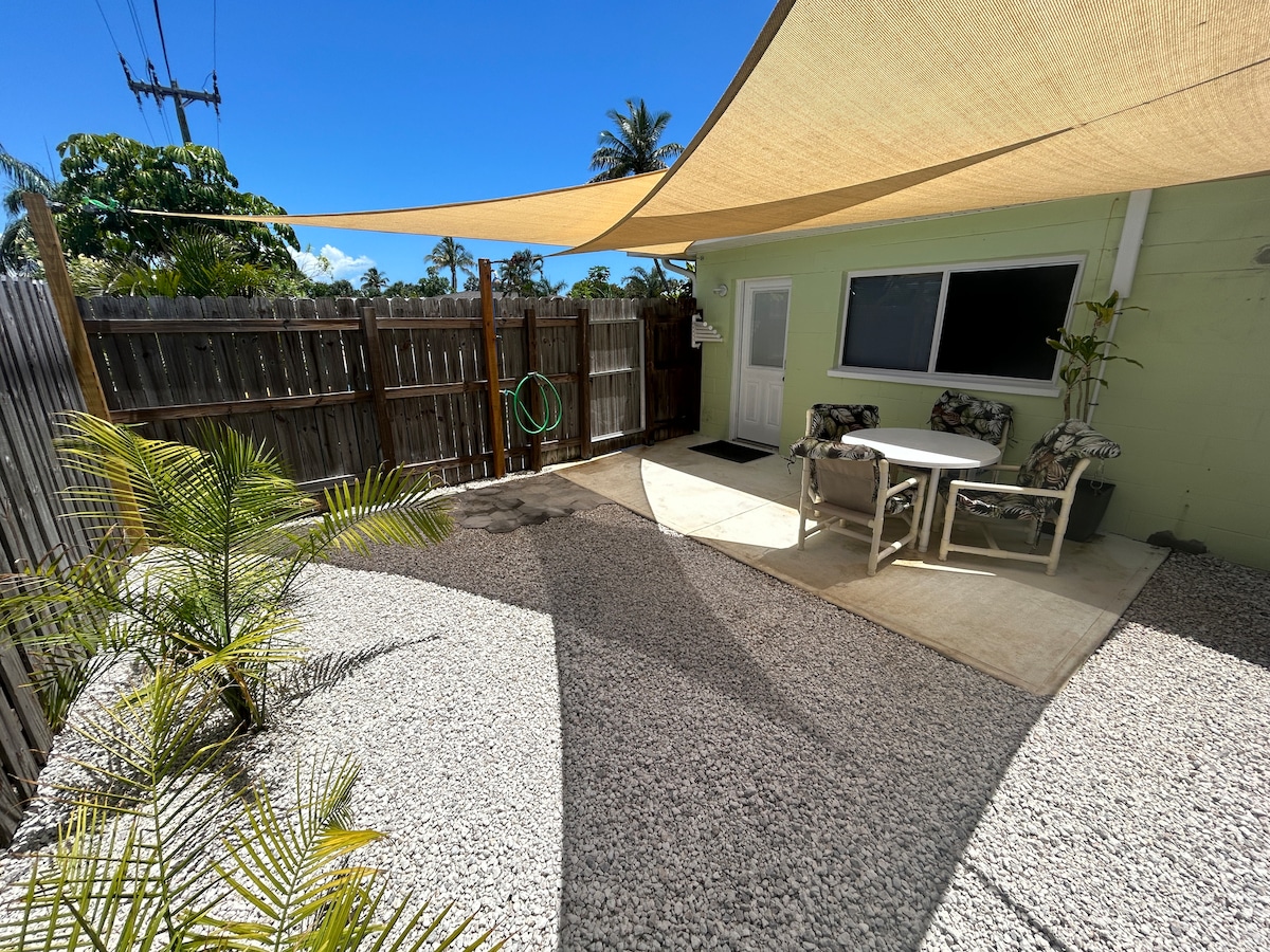 A shaded outdoor area features a small table surrounded by two chairs. Light colored gravel covers the ground, complemented by a subtle green wall and a potted plant. A back door leads to the property, framed by a wooden fence and blue sky.