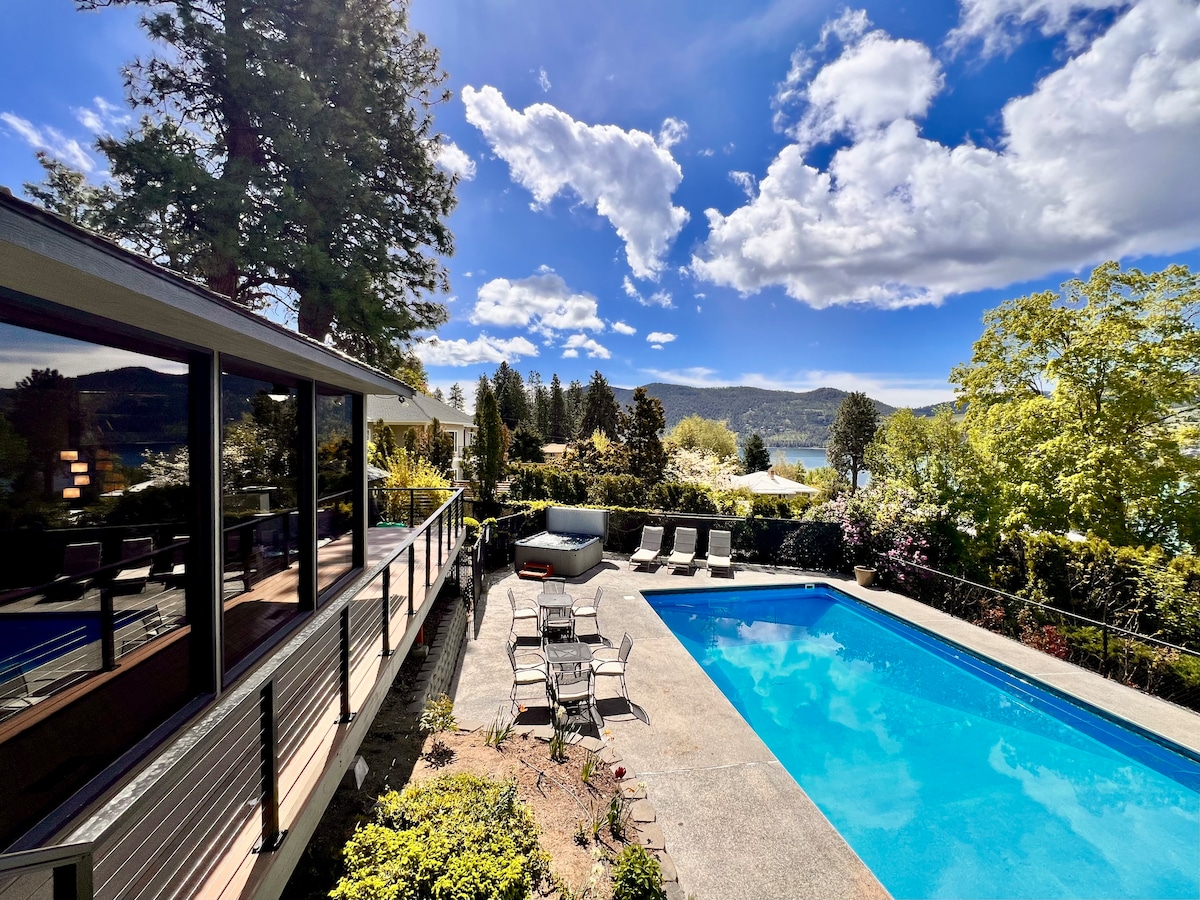 An expansive outdoor area features a spacious pool surrounded by a sun patio with lounge chairs and an outdoor dining set. Lush greenery and flowering plants border the space, with views of distant hills and a clear blue sky adorned with soft clouds.