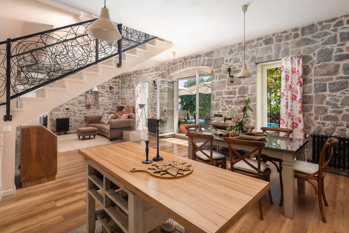 An open-concept living area is showcased, featuring a large wooden dining table with cross-back chairs. A comfortable seating area with a sofa and decorative pillows is visible. Natural light filters through large windows adorned with floral curtains, highlighting the stone walls and warm wooden flooring.