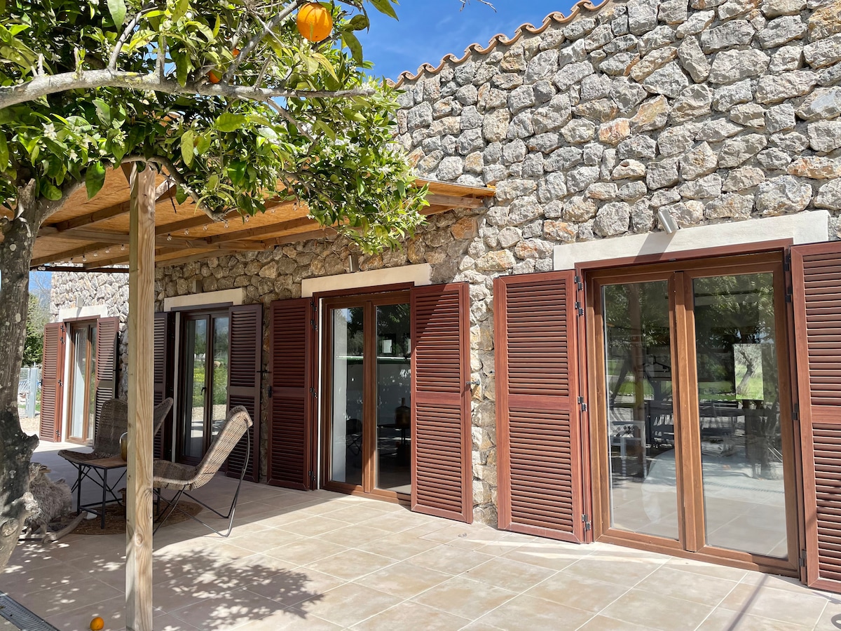 The exterior of the stone house is showcased with wooden shutters and a spacious covered terrace. A seating area with a chair is visible under the shade of a tree, with a backdrop of green fields and a clear blue sky.