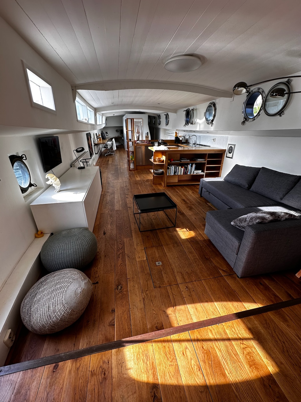 A spacious living area features wooden flooring and large windows allowing natural light to fill the room. A comfortable gray sofa faces a minimalistic coffee table, complemented by decorative poufs. Floating shelves hold books and decorative items, enhancing the open and airy feel.
