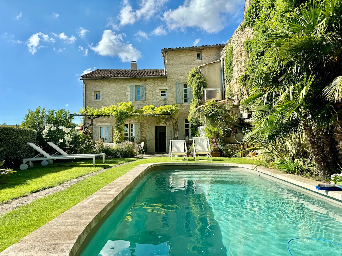 A beautifully landscaped garden surrounds a clear swimming pool, while lounge chairs are positioned on the lush green lawn. The stone house, showing a blend of rustic charm and modern comfort, features a variety of plants and trees, complementing the serene atmosphere.