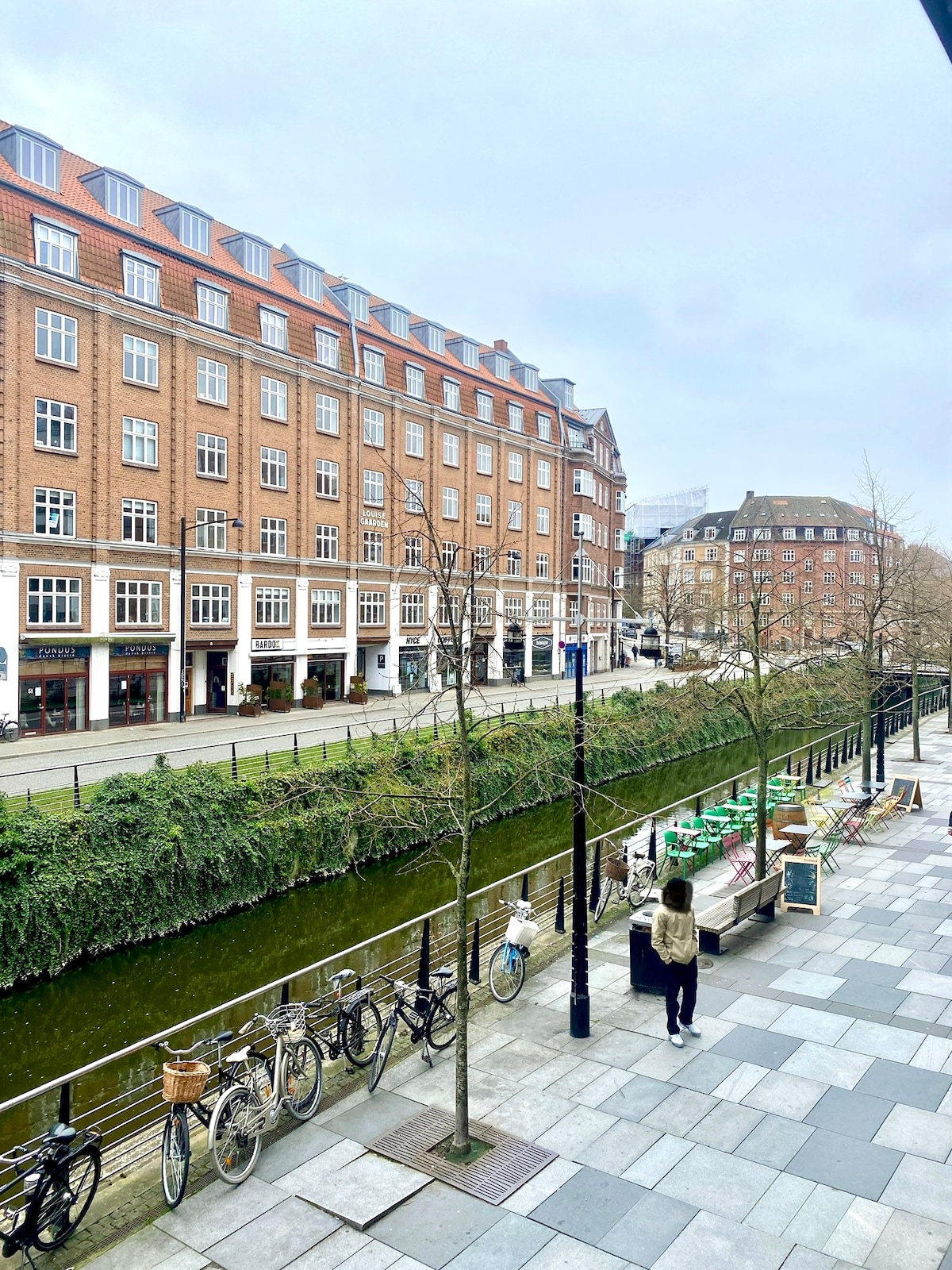 A view of a tree-lined street along a canal, featuring brick buildings with multiple windows. Bicycles are parked along the walkway, and a person can be seen strolling by. The ambiance suggests a vibrant urban setting, with cafes and shops visible in the distance.