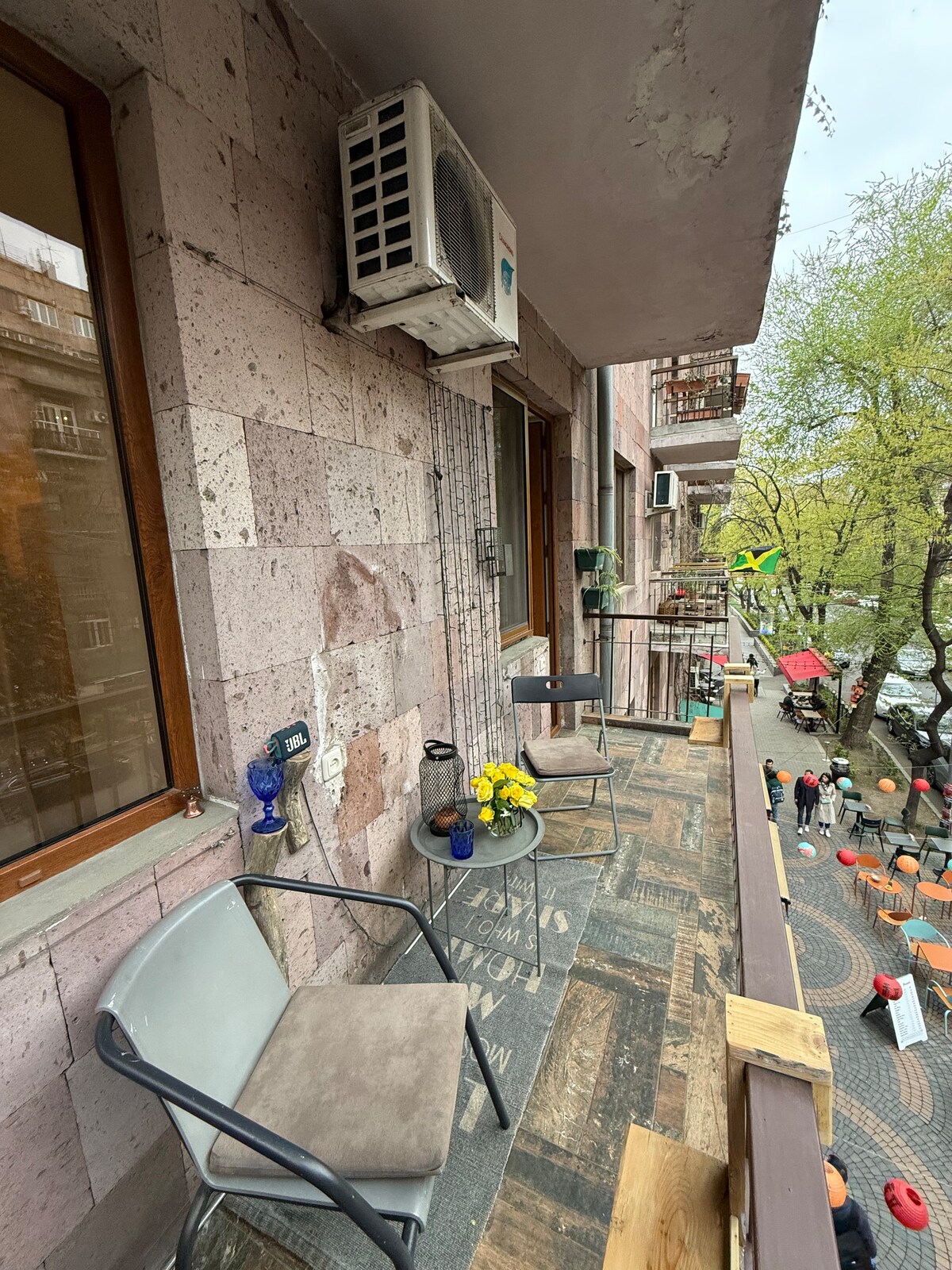 A small balcony is displayed, featuring two chairs and a glass table. Potted flowers add a pop of color. The building's stone facade is visible, along with neighboring balconies and greenery from nearby trees. The street scene includes hints of people enjoying the vibrant atmosphere below.