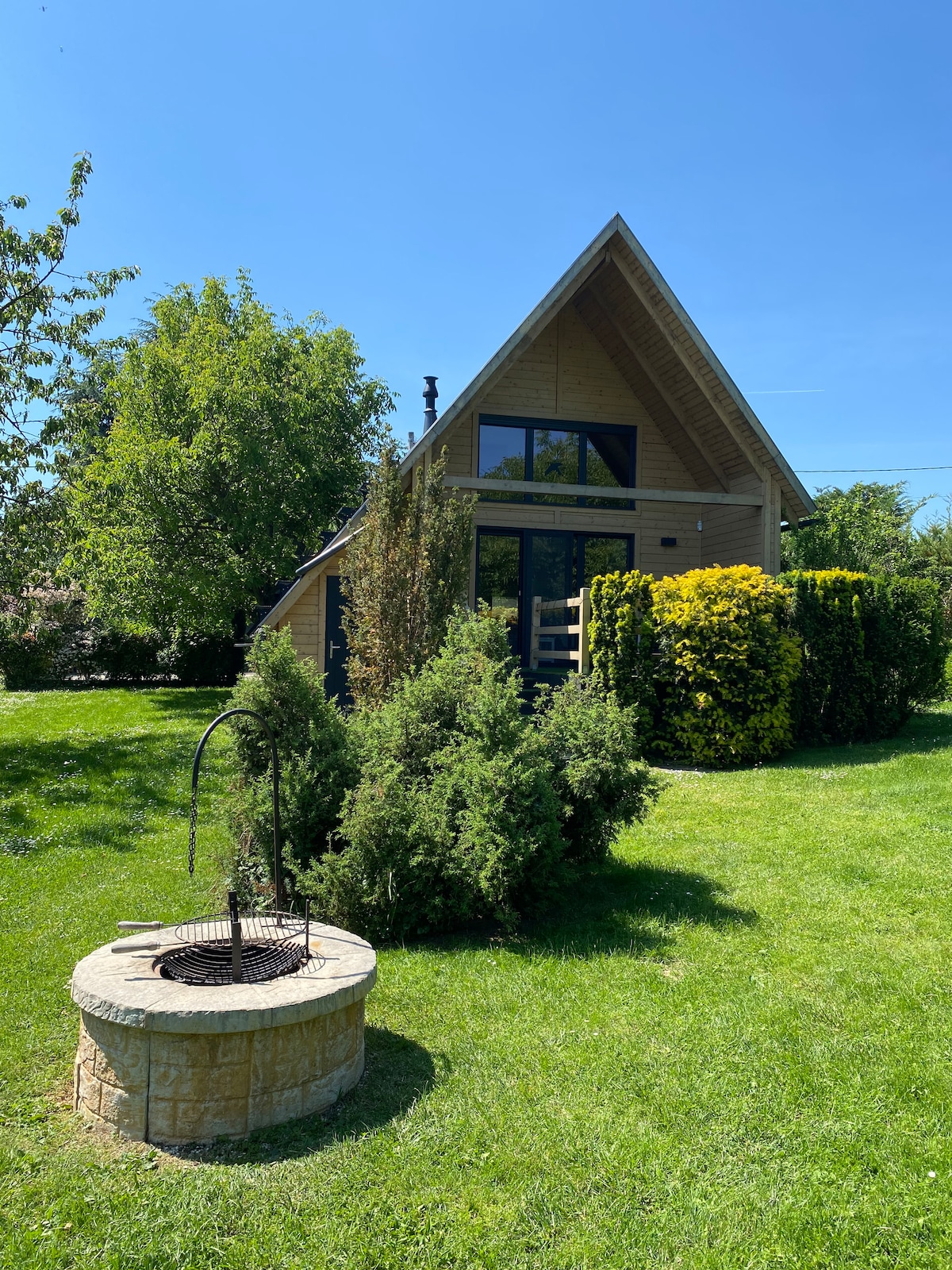 A modern wooden cabin is surrounded by lush greenery in a well-maintained garden. A stone well is situated in the foreground, complementing the natural landscape. Large windows reflect the clear blue sky, enhancing the serene outdoor setting.