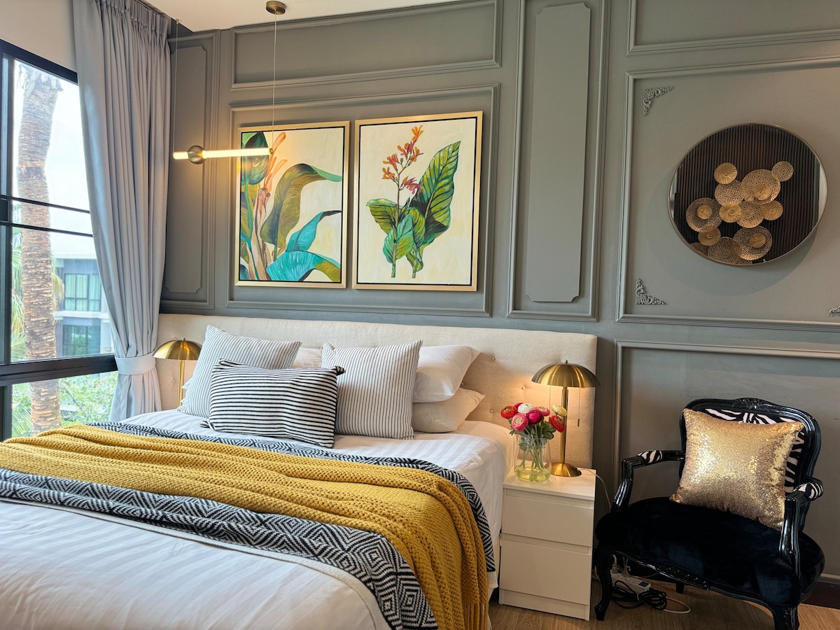 The bedroom features a spacious bed adorned with a textured yellow throw and patterned black and white pillows. Elegant bedside lamps illuminate the area, while framed botanical artwork decorates the wall. A stylish chair in the corner adds to the room's charm, complemented by a window showcasing greenery outside.