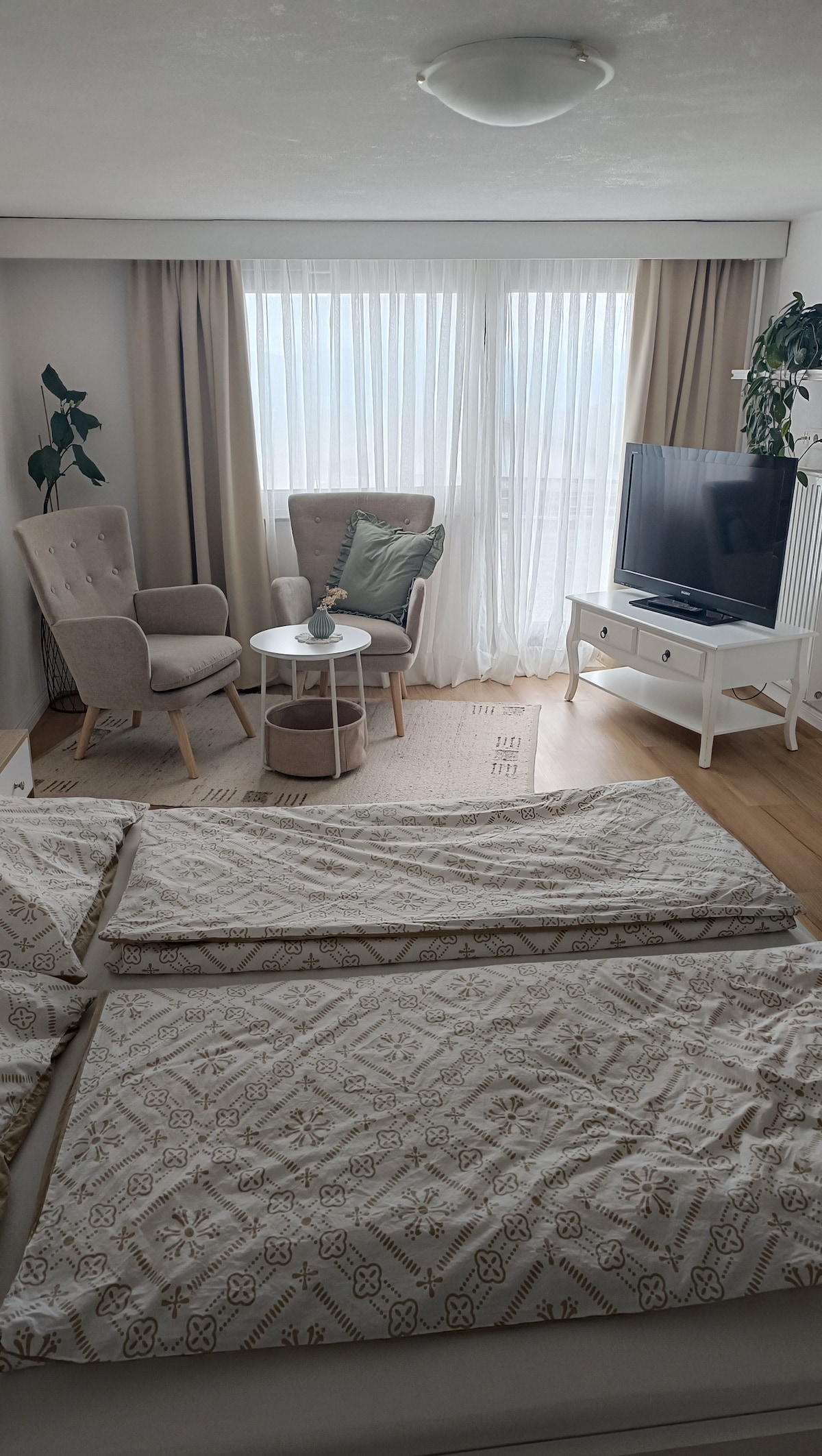 A bright bedroom features a spacious bed with an intricately patterned duvet cover. A cozy armchair sits next to a small table, creating a relaxing corner. Large windows allow natural light to fill the room, complemented by soft curtains and a neutral color palette.