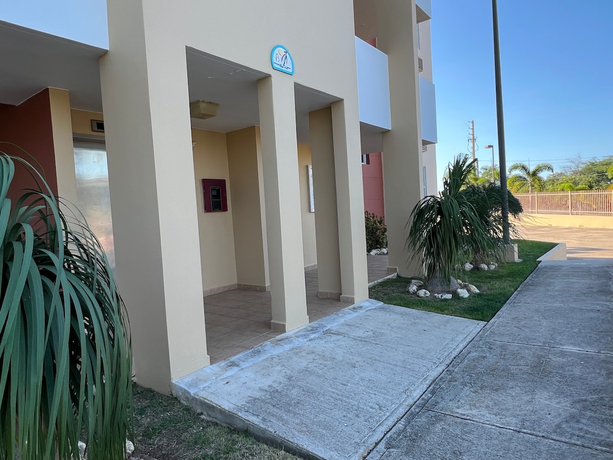 The entrance area of the building is displayed, featuring a tiled floor and bordered by greenery. A pathway leads to the door, which is marked with an address sign. The exterior walls are painted in warm tones, providing a welcoming appearance.