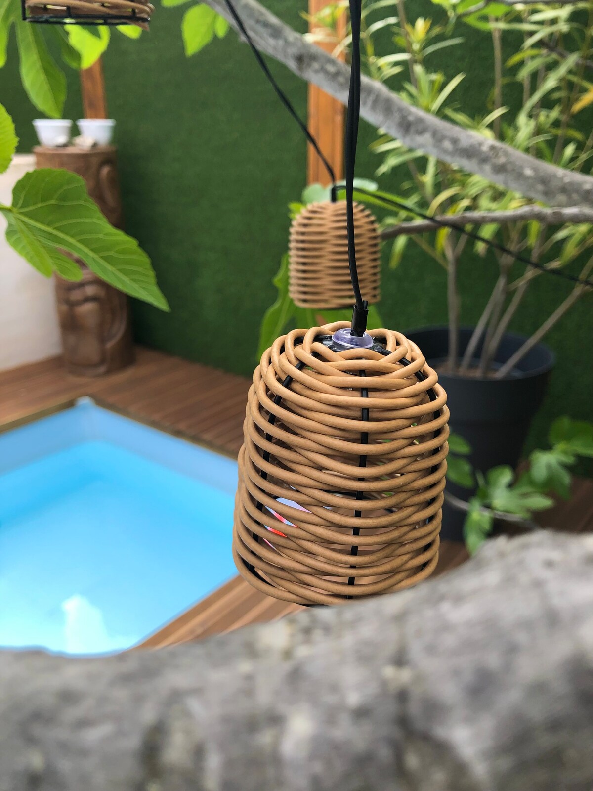 A woven rattan pendant light hangs from a tree branch, casting gentle shadows. The background features a small, inviting blue pool surrounded by wooden decking and greenery, adding a tranquil touch to the outdoor space.