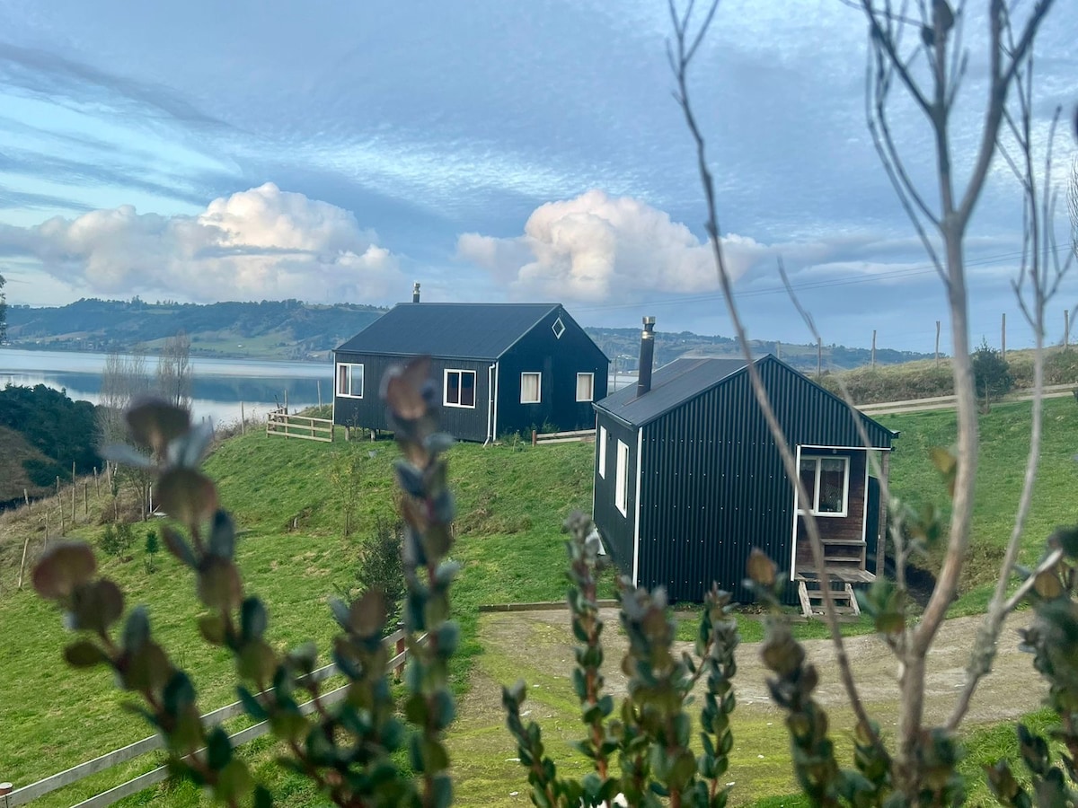 Two charming cabins are situated on a gently sloping green landscape, surrounded by tall grass and shrubs. In the background, a serene body of water reflects the sky, while soft clouds drift above. The cabins are designed with simple, rustic architecture.