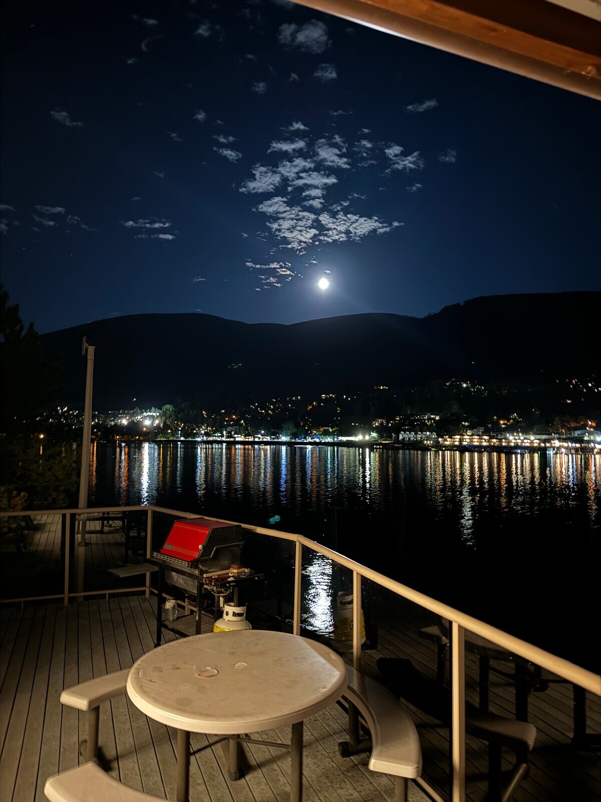 A serene outdoor deck is illuminated by moonlight, showcasing a circular table and a grill. Reflections of city lights and mountains can be seen on the calm water, creating a tranquil atmosphere. The night sky is partly cloudy, enhancing the peaceful setting.