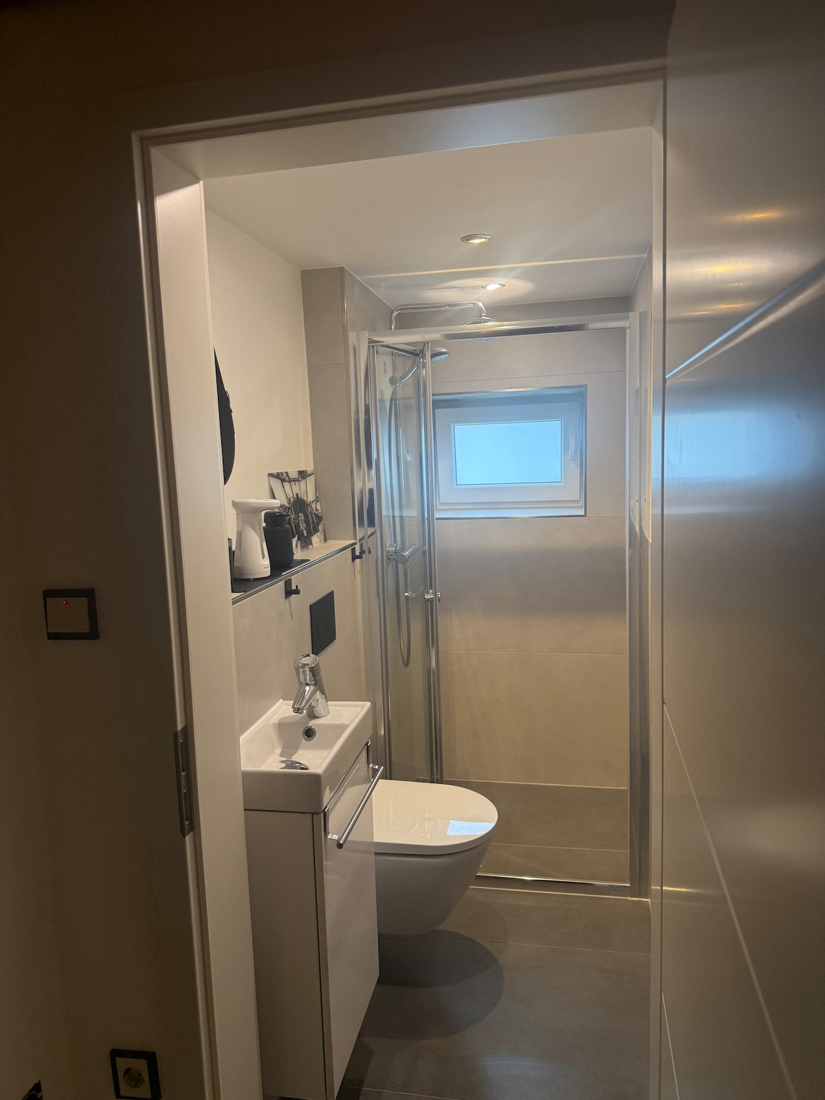 A modern bathroom is visible, featuring a sleek shower enclosed by glass. A compact white sink with a storage unit is positioned beside a toilet. Neutral-toned walls and floor tiles create a cohesive look, with natural light entering through a small window.