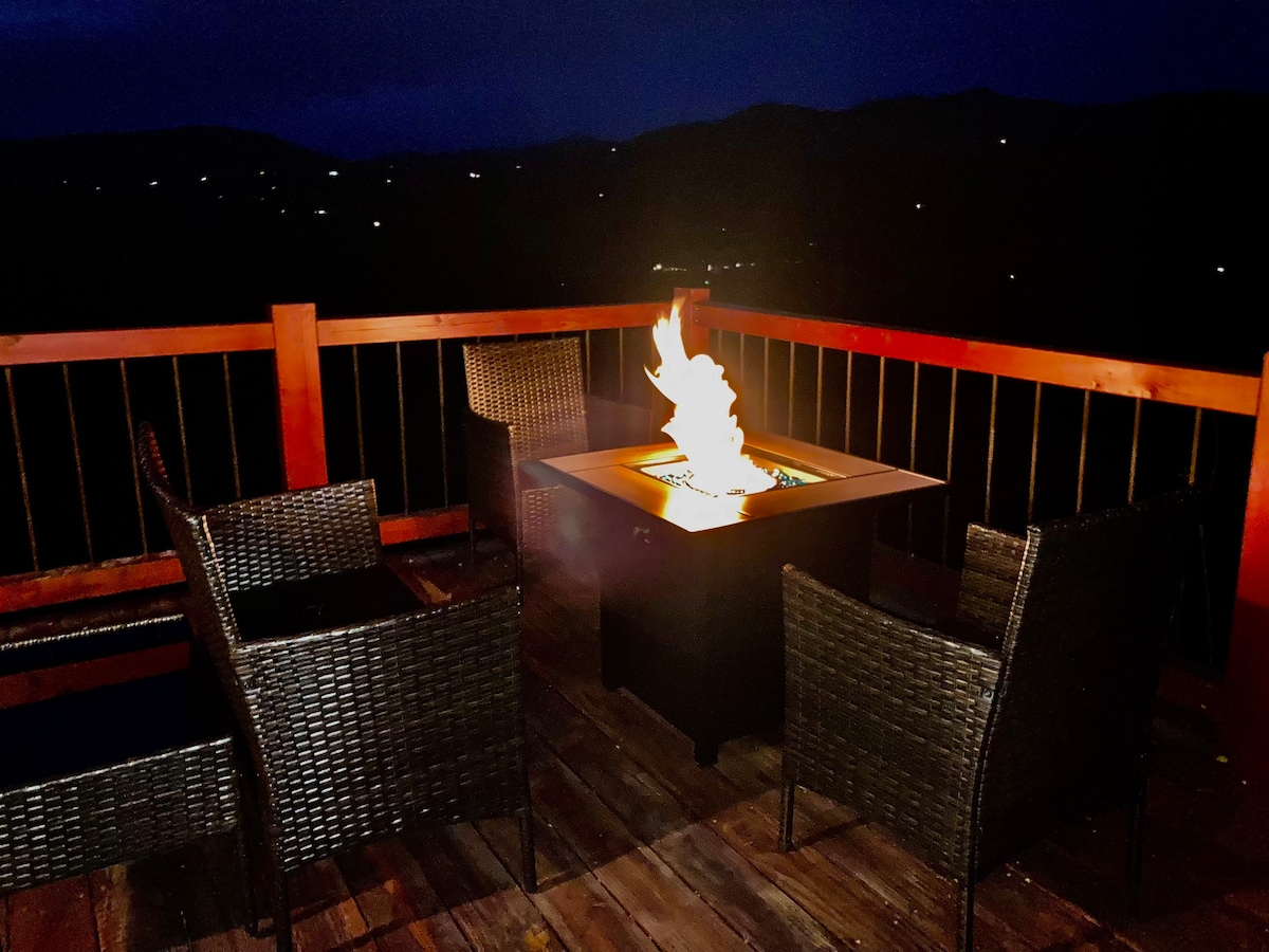 An outdoor patio features a fire pit surrounded by four wicker chairs, creating a cozy space for evening gatherings. Soft flames illuminate the area, while the surrounding landscape and distant lights from the valley are visible in the background.