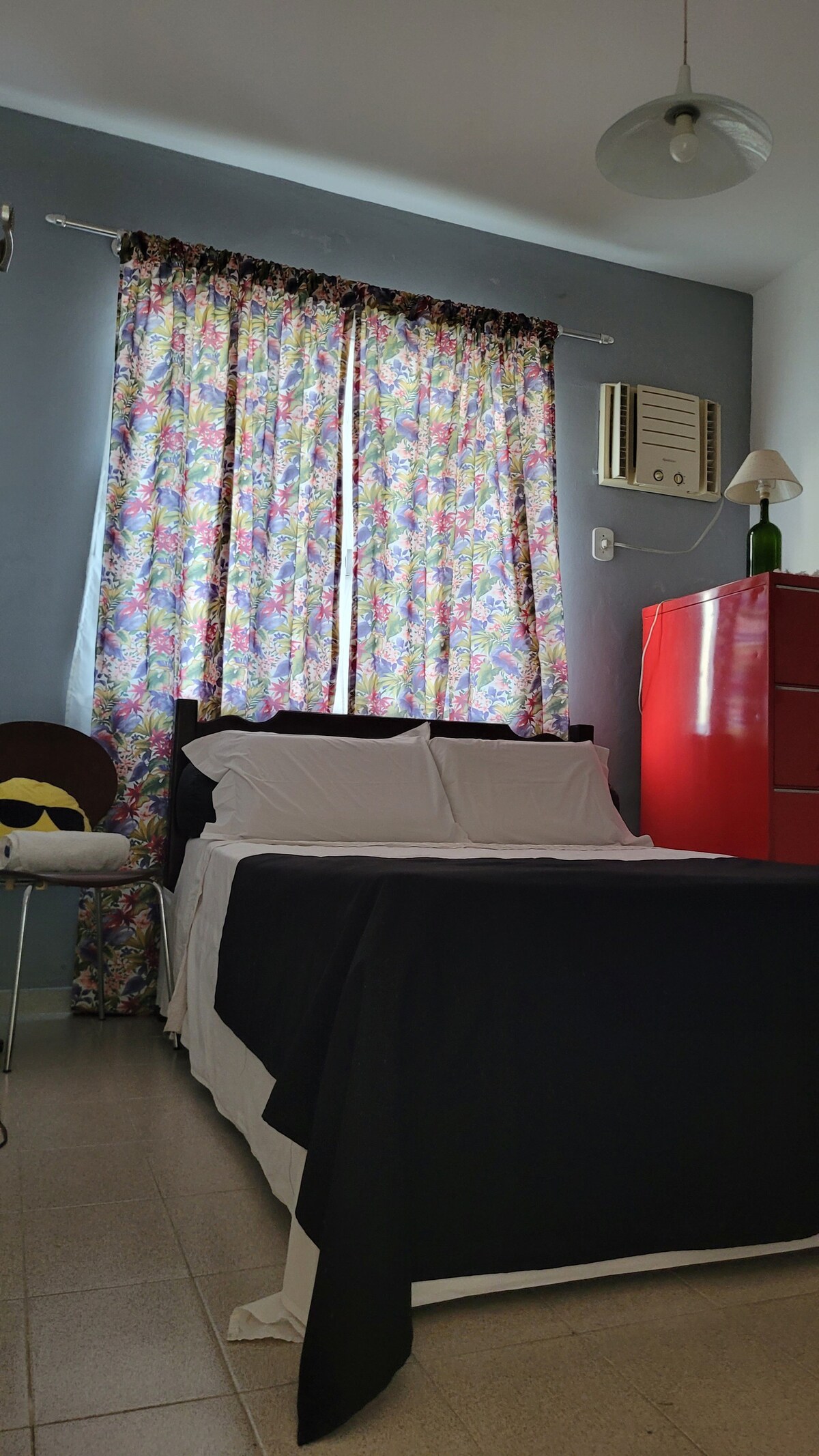 A cozy bedroom is equipped with a spacious bed adorned with white linens and a black bedspread. Colorful floral curtains frame a window, allowing natural light to illuminate the space. A red cabinet and a small lamp are positioned near the bed, contributing to the room's functionality.