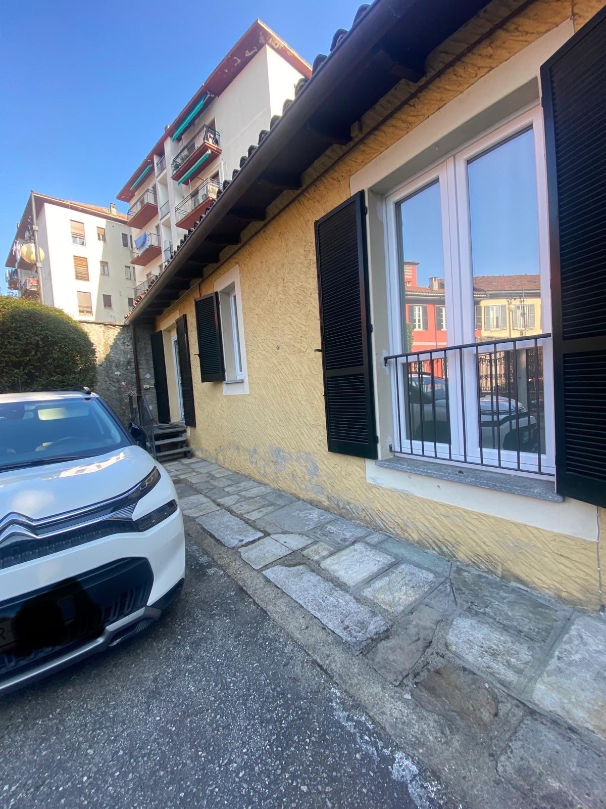 The exterior of the building features a yellow facade with black shutters on the windows. A well-maintained stone pathway leads to the entrance. A parked white car is visible in front, with multi-story buildings in the background, providing an urban context.