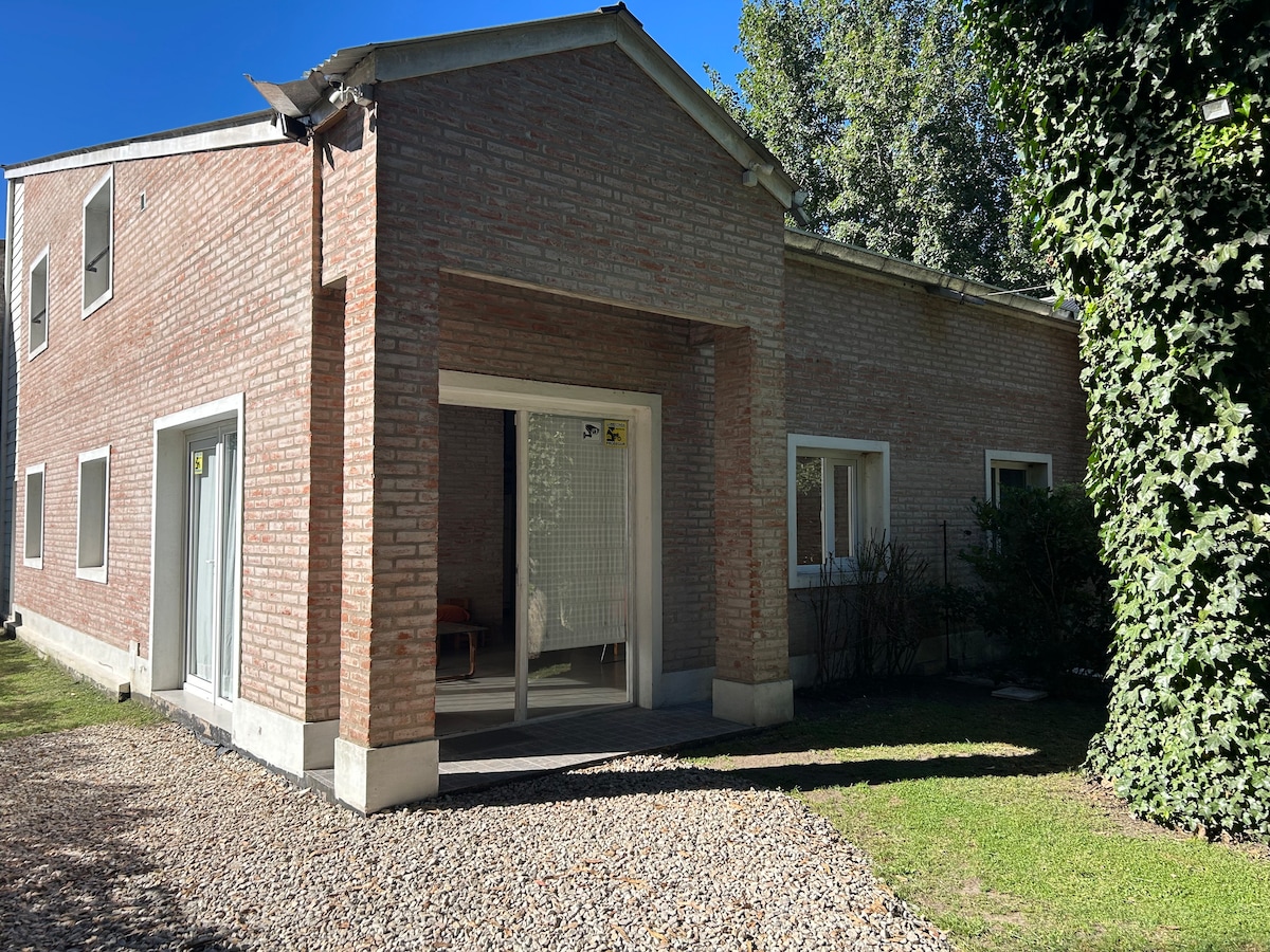 The exterior of the duplex is presented, showcasing a modern brick facade. Lush greenery surrounds the entrance, complemented by a well-maintained lawn. Large windows allow natural light into the interior, while a gravel pathway leads toward the entrance.
