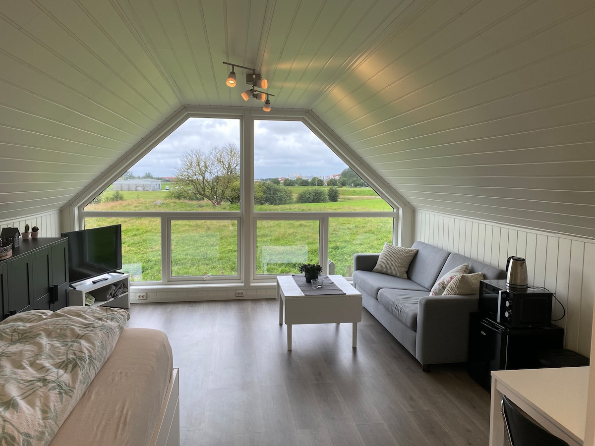 The bright living area features a gray sofa positioned beside a white coffee table. Panoramic windows provide expansive views of the surrounding greenery. A small kitchenette with a kettle and mini-fridge is visible towards the right, contributing to the cozy yet functional space.
