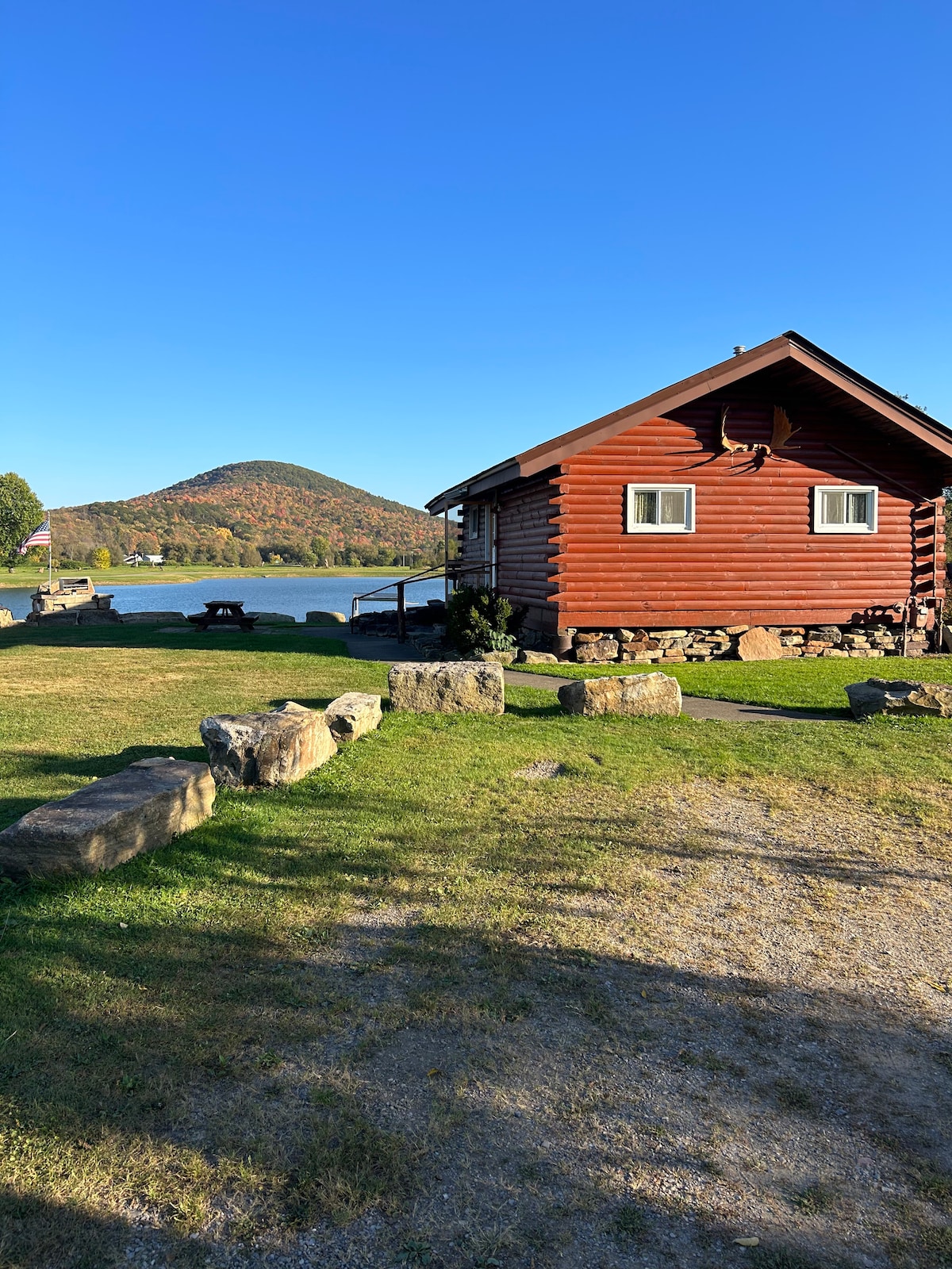 A rustic cabin is positioned by a serene lake, framed by lush greenery. Rolling hills rise in the background under a clear blue sky. The well-maintained lawn features natural stone borders, inviting outdoor relaxation and exploration along the water's edge.