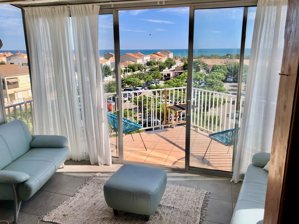 A bright living area features large glass sliding doors that open to a terrace with a view of the sea. Soft seating and a pouf are arranged on a textured rug, while sheer curtains gently frame the space, allowing natural light to enhance the inviting environment.