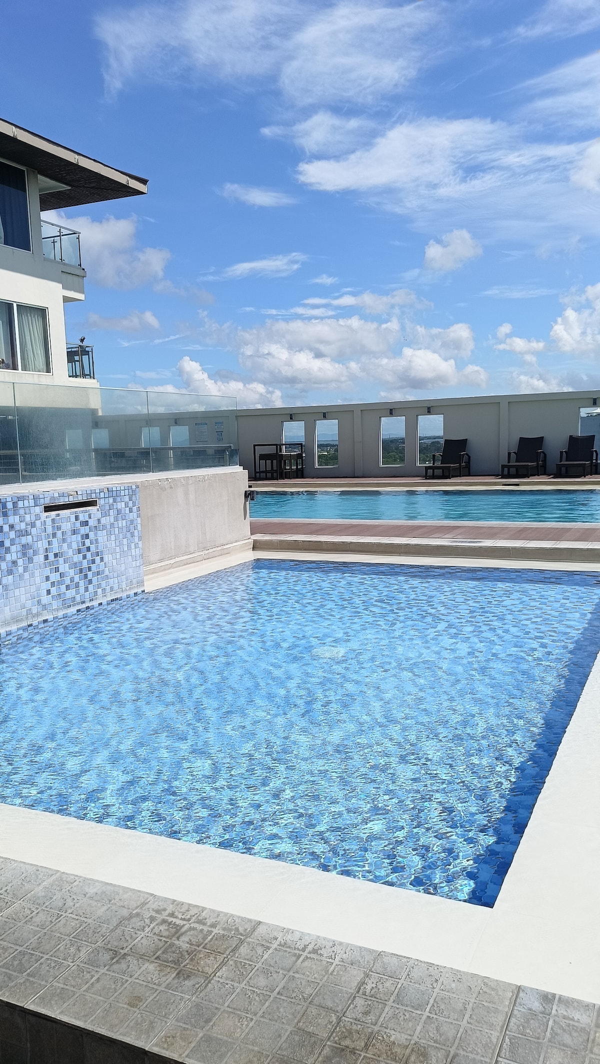 Two swimming pools are featured on a rooftop terrace, surrounded by light-colored tiles. One pool has a tranquil waterfall feature, while lounge chairs are positioned along the pool area. The bright blue sky and scattered clouds provide a picturesque backdrop.