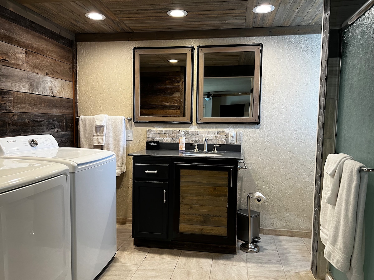 A well-appointed bathroom features a washer and dryer side by side, complemented by a sleek vanity with two mirrors and a contemporary faucet. Light-colored tiles cover the floor, while wooden accents on the wall add a rustic touch. A glass shower enclosure is visible in the corner.