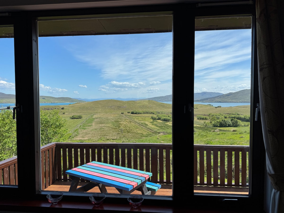 A large picture window frames sweeping views of the lush landscape, highlighting green fields and distant hills. A brightly colored picnic table is visible on the deck, inviting guests to enjoy outdoor dining in a serene setting.