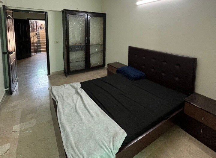 A spacious bedroom features a large bed covered with a black sheet and a white blanket draped casually at the foot. Two bedside tables are positioned beside the bed. A wooden wardrobe with glass doors stands against the wall, and a doorway leads to a staircase in the background.