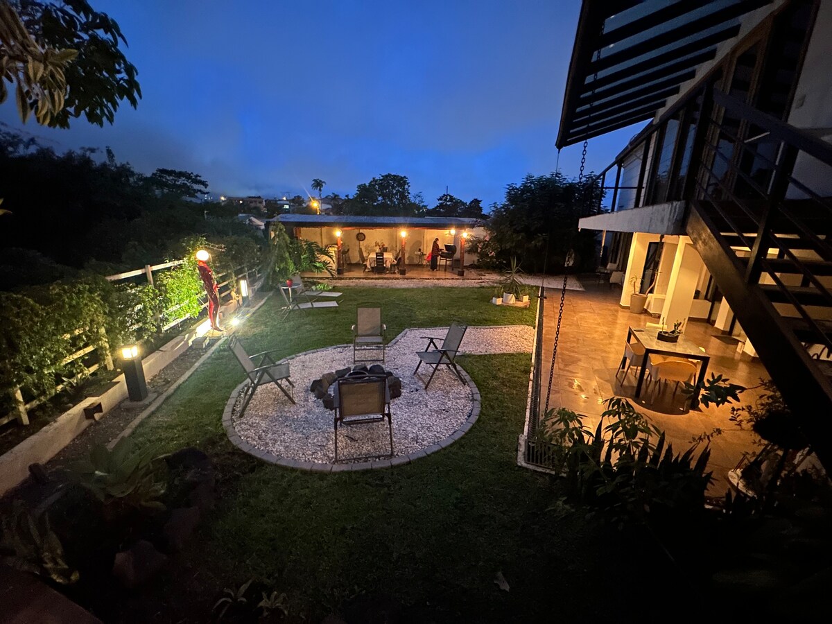 An outdoor space is visible at dusk, with cozy seating arranged around a central circular area of gravel. Soft lighting highlights the patio area, creating an inviting environment, while lush greenery surrounds the space, contributing to a serene atmosphere for relaxation.