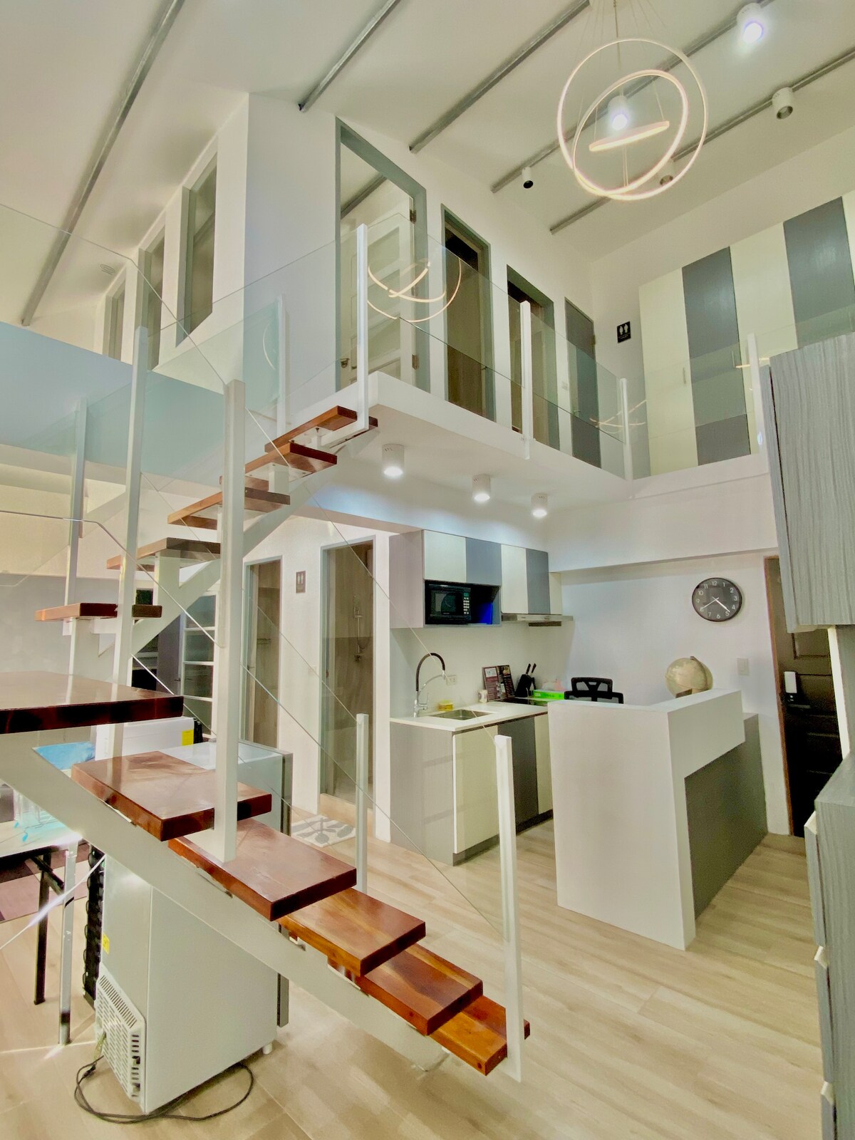 An open-plan area features a modern staircase with wooden steps and glass rails leading to a lofted section. A well-equipped kitchen is visible with sleek cabinetry and a circular wall clock. Soft lighting enhances the spacious feel of the living area.