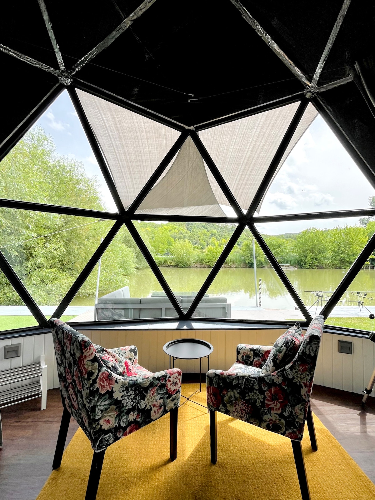 Two floral upholstered chairs are positioned around a small black table, offering a relaxing spot near large triangular windows. The view outside reveals a serene private lake surrounded by lush greenery, allowing natural light to fill the space.