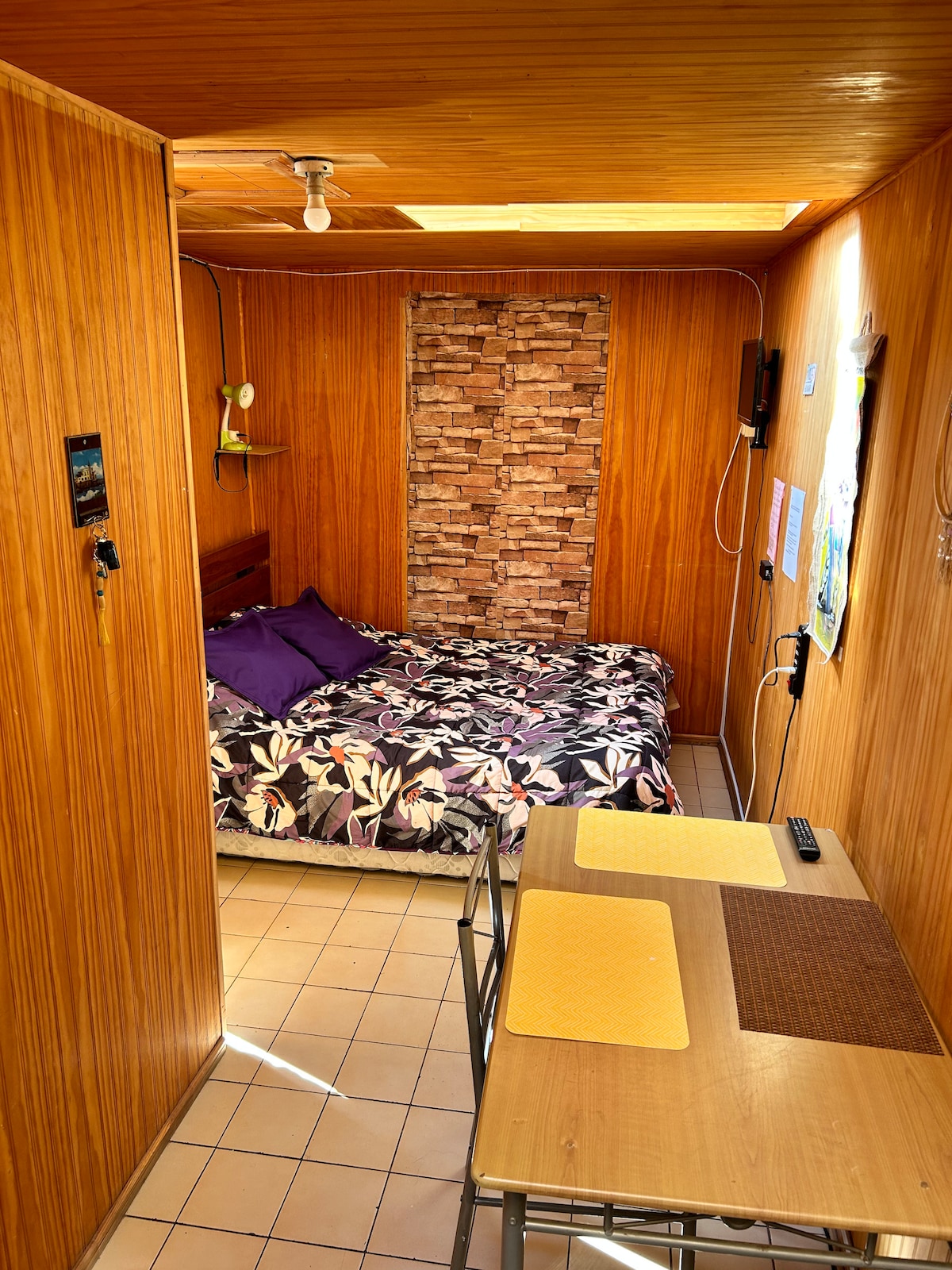 A cozy room features wood paneling and tiled flooring, with a spacious bed dressed in a floral bedspread. A small desk area offers two chairs, and a stone accent wall adds character. Natural light enters through a window, illuminating the warm space.