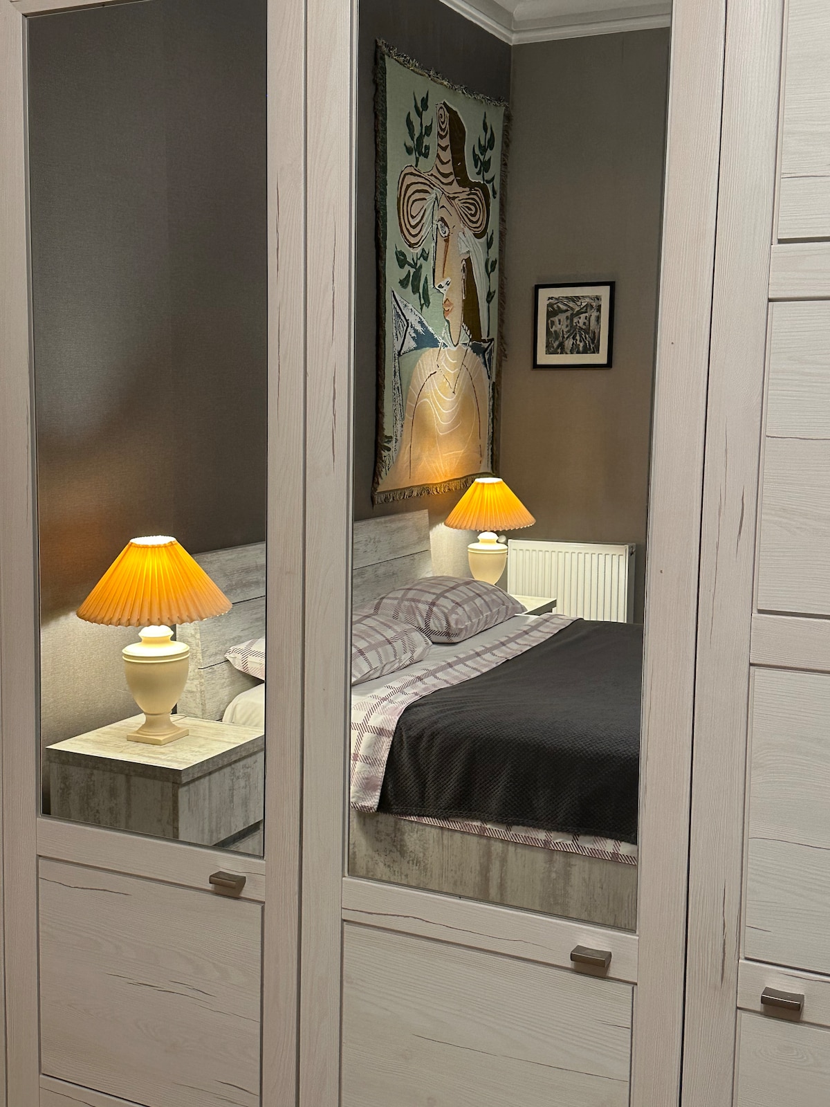 A warmly lit bedroom is reflected in a large mirror. The space features a spacious bed with a dark bedding set, complemented by soft gray pillows. Two lamps illuminate the bedside table, while a decorative wall hanging adds an artistic touch to the room's ambiance.