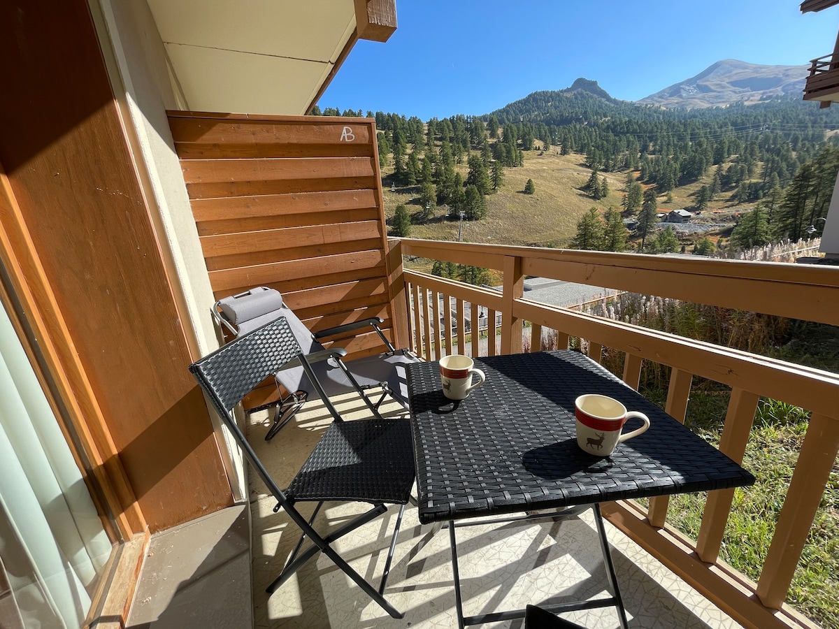 A balcony is shown, featuring a small table with two cups and two folding chairs. A sun lounger is positioned nearby. The surrounding landscape of mountains and greenery is visible, providing a tranquil outdoor setting.