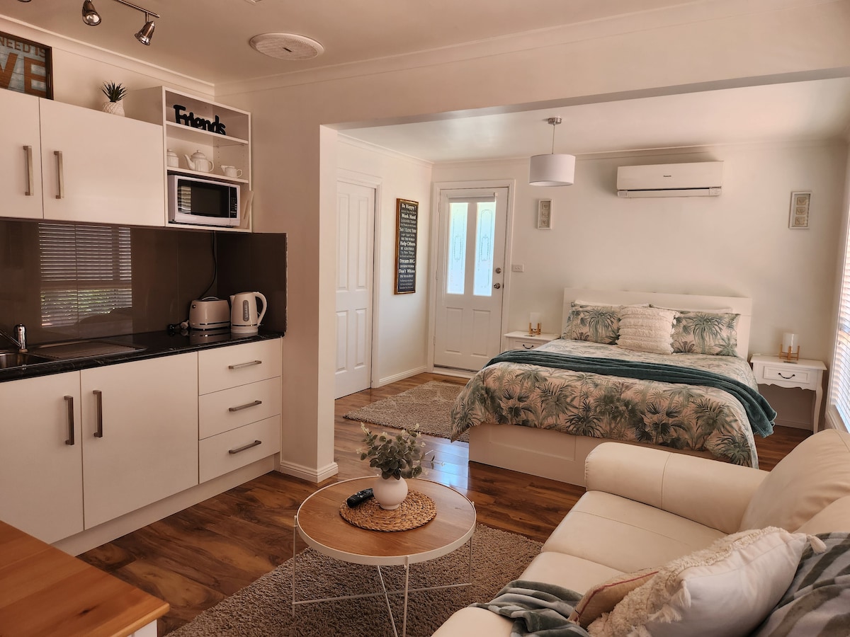 A self-contained studio apartment features a queen-size bed with a decorative quilt. A cozy lounge area includes a light-colored sofa and coffee table. The kitchenette is equipped with a microwave and kettle. Natural light enters through the entrance door, complementing the warm tones of the wood flooring.