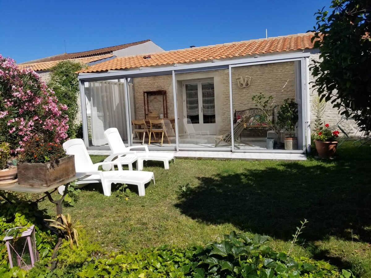 A spacious garden features a glassed-in veranda, which provides an open view of the interior. Two white lounge chairs are positioned on the lawn, surrounded by green grass and blooming flowers, creating a pleasant outdoor relaxation area.