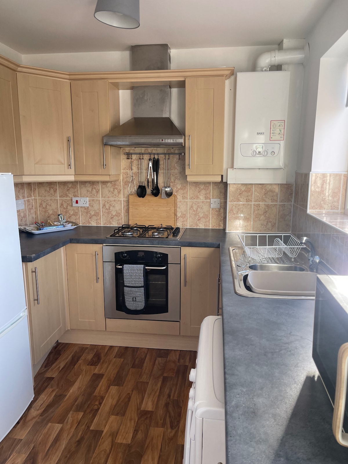The kitchen features wooden cabinetry with a modern design, equipped with a gas stove, oven, and stainless steel sink. A white refrigerator stands in the corner, and a window allows natural light to brighten the space, reflecting off the tiled backsplash.