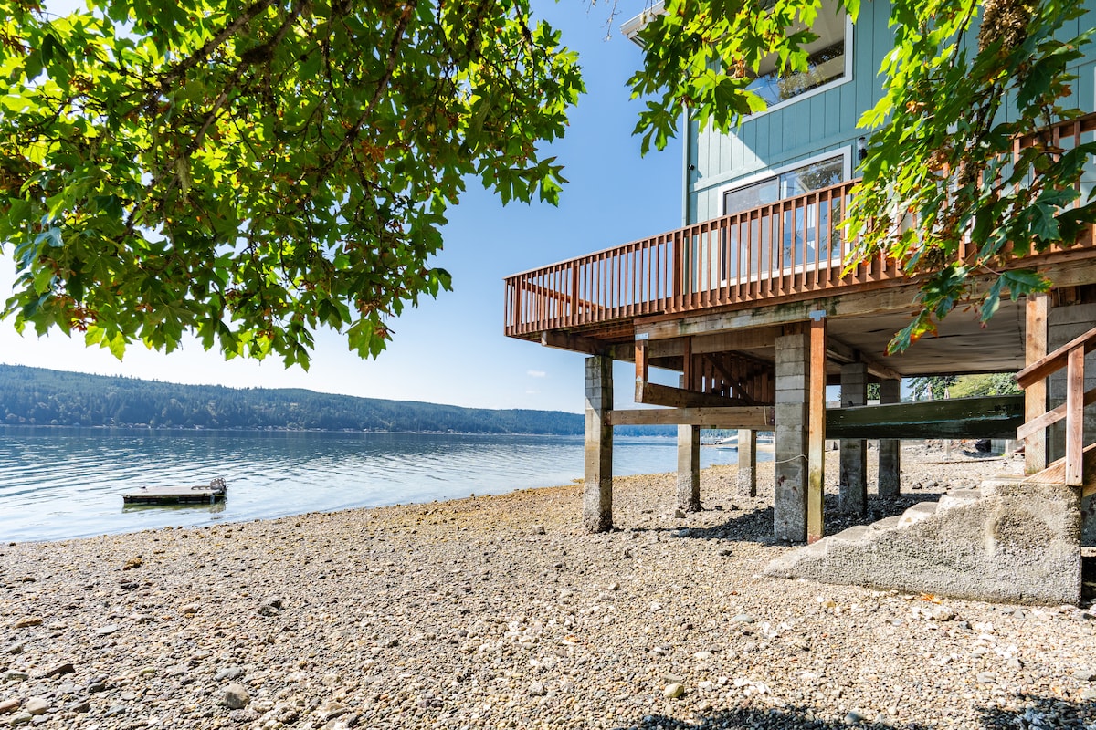 The house is elevated on stilts above a rocky beach, surrounded by a serene water view. Sunlight filters through leafy trees, casting gentle shadows on the patio area beneath the home, which leads to the calm surface of the Hood Canal.
