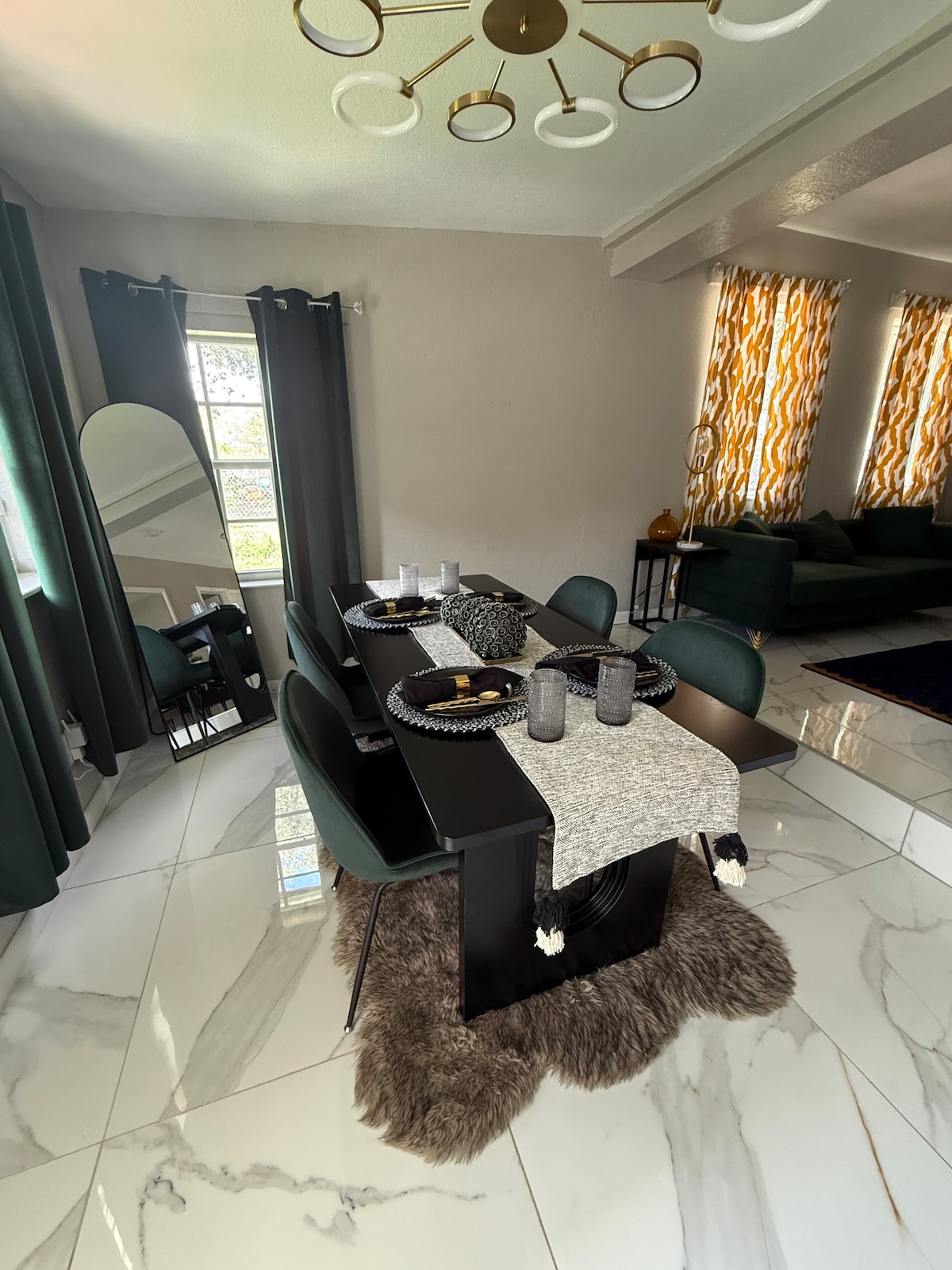 A dining area is presented with a sleek black table set for four. Modern black chairs surround the table, complemented by a decorative runner and tableware. Large windows adorned with patterned curtains allow natural light to brighten the space, while a plush rug adds warmth beneath the table.