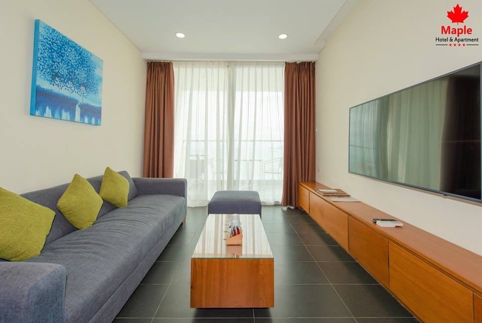 A modern living room features a large, gray sofa complemented by green cushions. A wooden coffee table sits in front, with a flat-screen television mounted on the wall opposite. The room is illuminated by natural light from the floor-to-ceiling curtains.