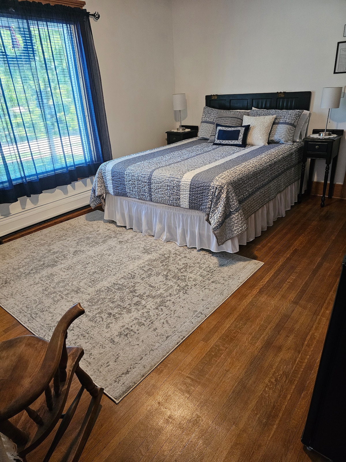 A cozy bedroom is shown, featuring a spacious bed with a blue and white patterned coverlet. Bedside tables are positioned on either side, with matching lamps. A large area rug in neutral tones lies beneath the bed, and natural light filters through a window covered by blue curtains.