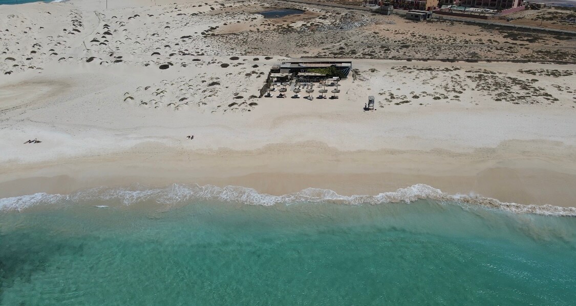 A serene beach scene is depicted, featuring soft white sand and crystal-clear turquoise water. Sun loungers are positioned under shade structures, providing comfort for relaxation. The coastline gently curves, merging with the horizon where it meets an expansive sky.