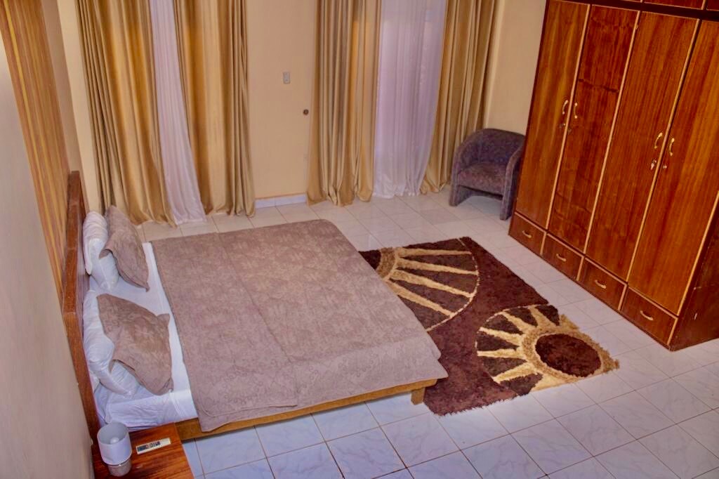 A spacious sleeping area features a large bed with soft bedding set on a textured area rug. Light-colored curtains frame the window, while warm-toned drapes add a touch of elegance. A wooden wardrobe stands against the wall, and a small upholstered chair is positioned nearby.