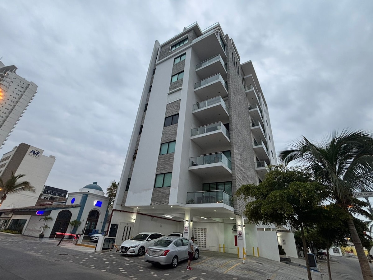 A modern five-story building features a combination of textured stone and sleek white facades. Balconies with glass railings provide outdoor spaces. Cars are parked in front, and palm trees are visible nearby, contributing to the urban landscape.