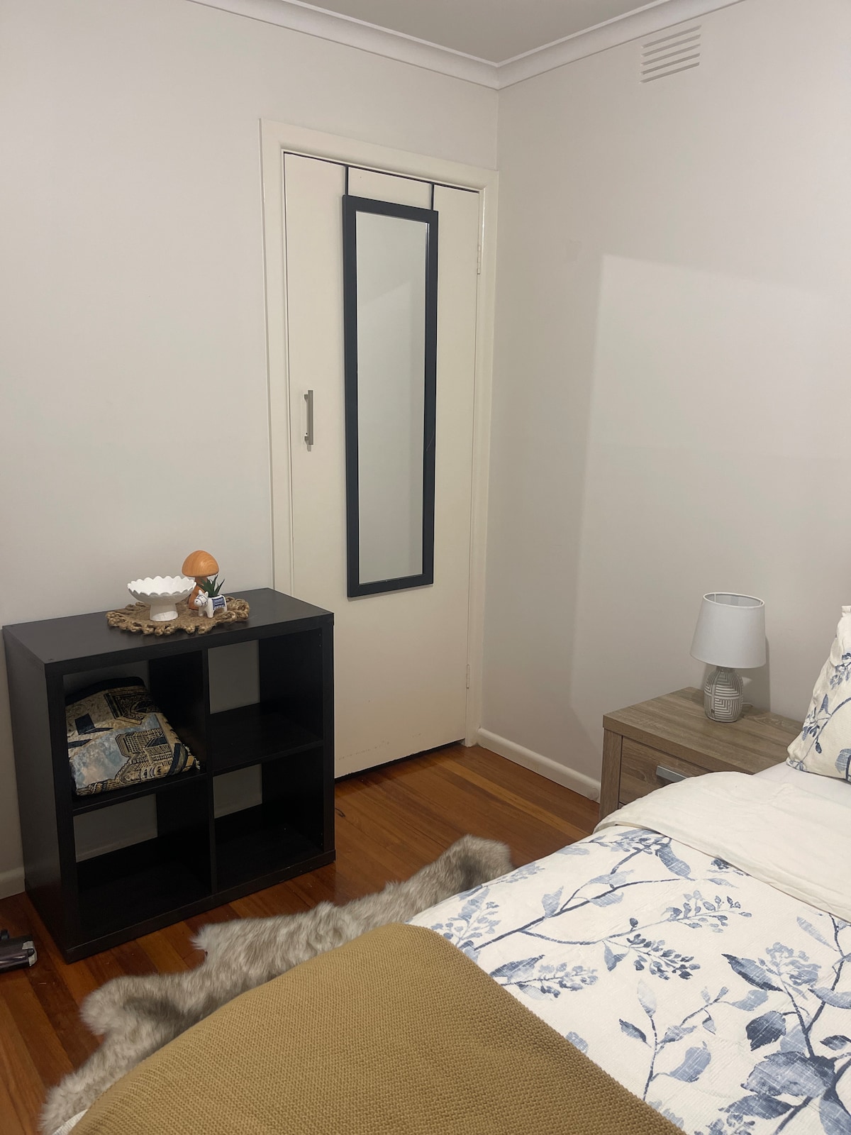 A neutral-toned bedroom features a bedside table with a lamp and decorative items. A fitted wardrobe is positioned against the wall, while a stylish shelving unit displays various items. Soft textures are present on the bed, complemented by a cozy blanket.