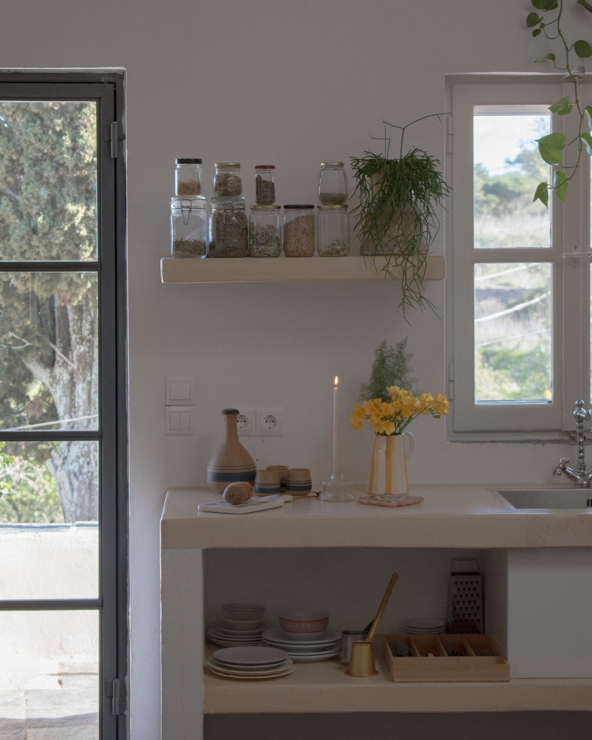 A clean and bright kitchen area features a small countertop with an array of spices stored in glass jars on a shelf. A candle and a vase of yellow flowers add a decorative touch, while a window provides natural light and views of the outdoors.