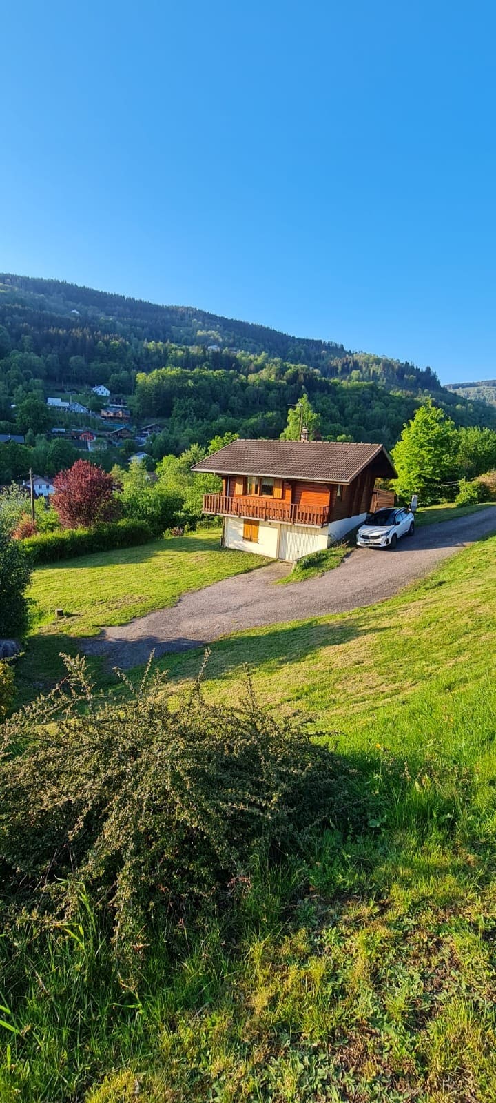 A charming chalet is positioned on a gentle slope, surrounded by lush greenery and trees. The building features a wooden exterior, a balcony, and large windows. A driveway leads up to the garage, displaying a parked car, with distant mountains visible in the background under a clear blue sky.