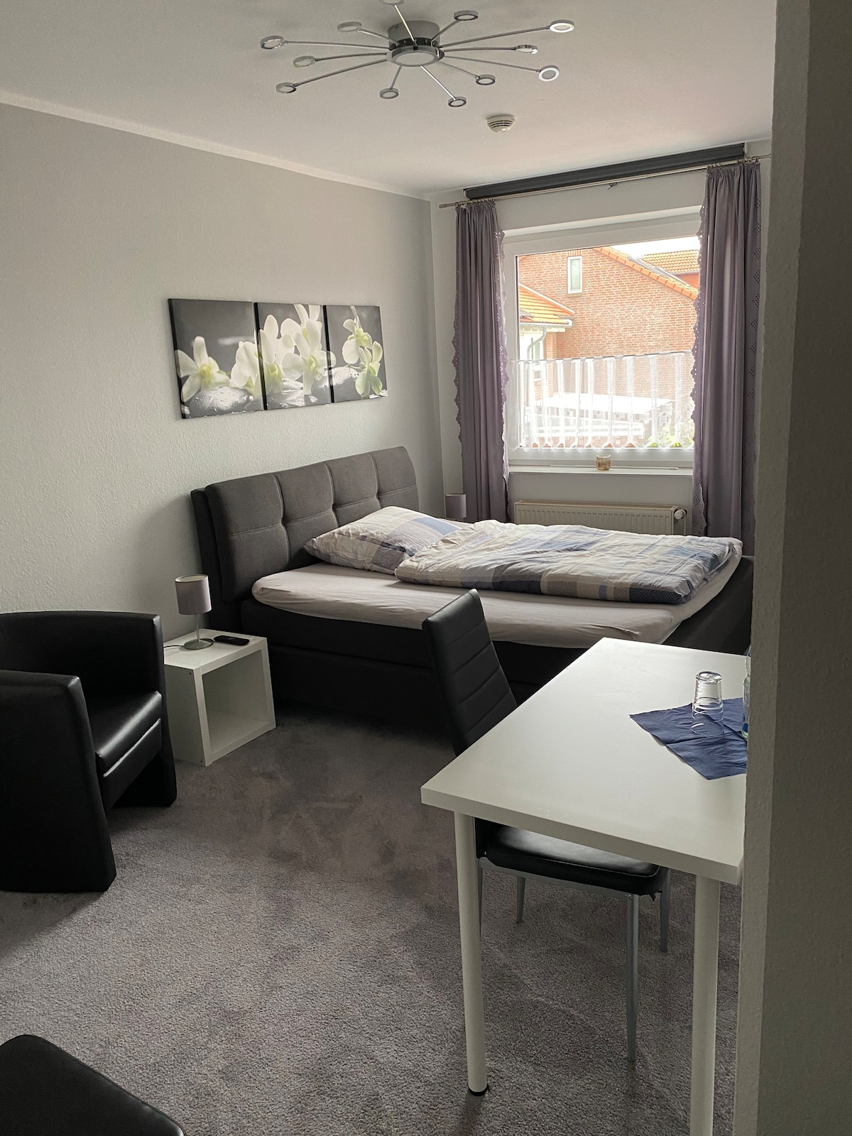 A compact bedroom features a spacious bed with a checkered blanket, positioned near a window draped with sheer curtains. Two black chairs and a small table are arranged in the room, complemented by a modern light fixture on the ceiling. Wall art depicting flowers adds a touch of color.